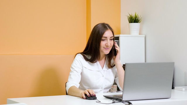 smiling lady receptionist in white coat talks on smartphone explaining to customer price list at laptop in beauty salon, copy space
