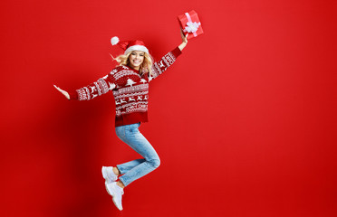 happy young cheerful girl laughs and jumps in christmas hat and with  gift on  red   background