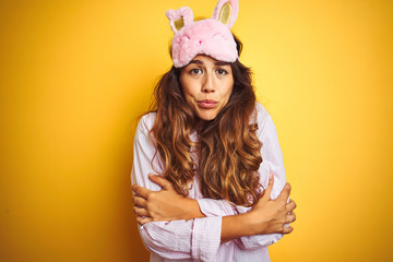 Young woman wearing pajama and sleep mask standing over yellow isolated background shaking and...