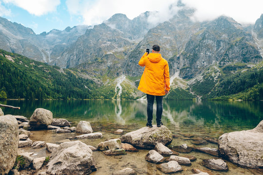 man in yellow raincoat taking picture on phone of lake in mountains