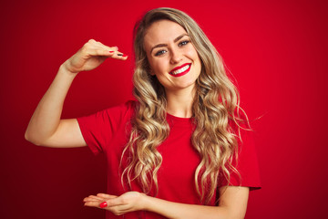 Fototapeta premium Young beautiful woman wearing basic t-shirt standing over red isolated background gesturing with hands showing big and large size sign, measure symbol. Smiling looking at the camera