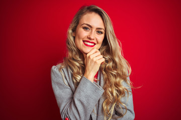 Young beautiful business woman wearing elegant jacket standing over red isolated background looking confident at the camera with smile with crossed arms and hand raised on chin. Thinking positive.