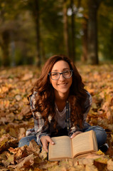 Beautiful girl in glasses sitting on the ground and reading book in park. Leisure time on warm autumn day. Autumn mood, enjoy the season