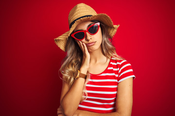 Young beautiful woman wearing sunglasses and summer hat over red isolated background thinking looking tired and bored with depression problems with crossed arms.