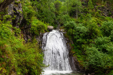 A large waterfall in the back of the Altai Mountains with gray-brown stones near a steep cliff with green trees. Rest and loneliness while traveling to deserted places.