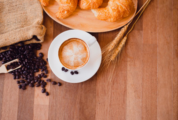 Top view white hot coffee cup and beans on wooden table with croissant bread background.Latte art coffee menu for breakfast in the coffee shop.Selective focus.