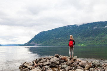 View from the back of a young beautiful blonde woman who is standing on the rocky shore of Lake Teletskoye with a red backpack during an excursion in the Altai mountains. Travel and tourism.