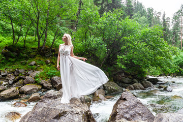 A young blonde girl in an elegant pose pulls up a boudoir dress in the mountains against a waterfall and stones raising her hands up like a ballerina on a warm summer day in Altai with green trees