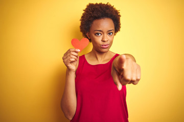 African american woman holding romantic paper hearts over yellow isolated background pointing with finger to the camera and to you, hand sign, positive and confident gesture from the front