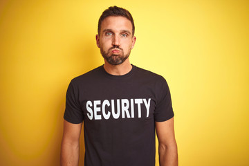 Young safeguard man wearing security uniform over yellow isolated background puffing cheeks with funny face. Mouth inflated with air, crazy expression.
