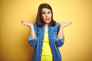 Young beautiful brunette woman wearing casual denim jacket over yellow isolated background clueless...