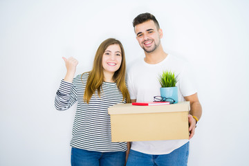 Young couple holding cardboard box moving to a new home over white isolated background pointing and showing with thumb up to the side with happy face smiling