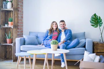 Young beautiful couple sitting and hugging on the sofa at new apartment with cardboard boxes around them, very excited for moving to a new home