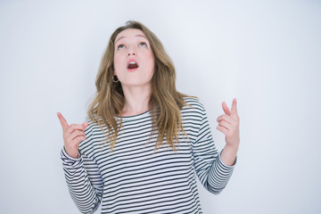 Beautiful blonde girl with blue eyes wearing striped sweater over white isolated background amazed and surprised looking up and pointing with fingers and raised arms.