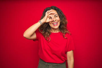 Middle age senior woman with curly hair over red isolated background doing ok gesture with hand smiling, eye looking through fingers with happy face.