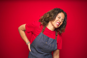 Middle age senior woman wearing apron uniform over red isolated background Suffering of backache, touching back with hand, muscular pain