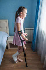 Girl child with a broken leg in a cast is standing on crutches and looks out the window