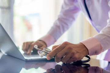 Male doctor,physician or practitioner hand holding stethoscope ,standing and work on laptop computer at office.Medic tech,teleconference,telemedicine,electronic,health or medical record system concept