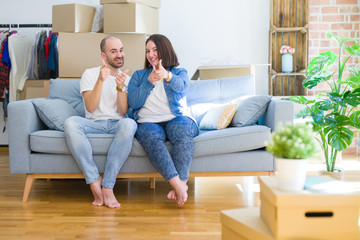 Fototapeta na wymiar Young couple sitting on the sofa arround cardboard boxes moving to a new house pointing fingers to camera with happy and funny face. Good energy and vibes.