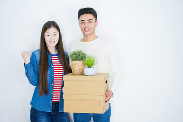 Beautiful young asian couple over white background holding cardboard boxes screaming proud and celebrating victory and success very excited, cheering emotion