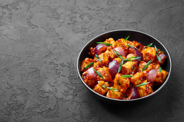 Schezwan Paneer in black bowl at dark slate background. Schezwan Paneer is indo-chinese cuisine dish with deep fried Paneer cheese, onion and Schezwan Sauce. Copy Space