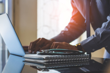 Smart male doctor ,hands typing on laptop computer keyboard with white notebook and medical stethoscope on the desk at clinic or hospital. Medic tech,medicare, telemedicine or teleconference concept.