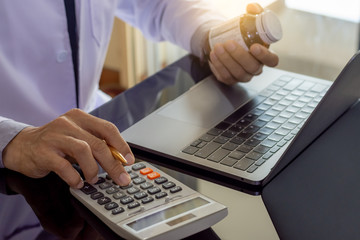 Male doctor in white lab coat,hand holding prescription medicine bottle and using calculator,work on laptop computer on the desk at office.
