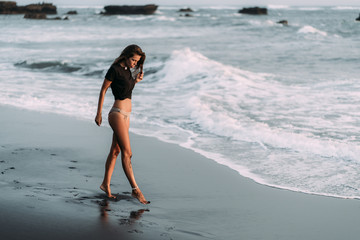 Fototapeta premium Girl with sexy body in black T-shirt and swimwear walks on black sandy beach