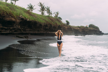 Beautiful girl in dark swimsuit walks on black sand beach near ocean