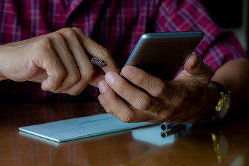 Closeup casual young business man hand using mobile smart phone ,plan to write and sign cheque book on the desk at office. Business technology and paycheck concept.