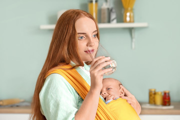 Young mother with little baby in sling drinking water at home