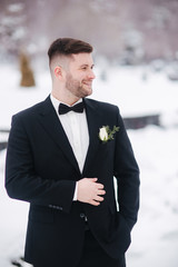 Handsome bearded man in black suit with tie bow walking in winter park. Young groom outside in winter