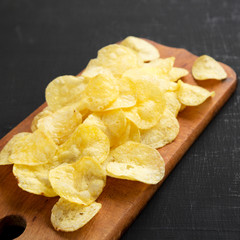 Yellow potato chips with salt on a rustic wooden board on a black surface, side view. Closeup.