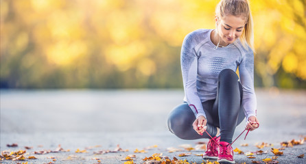 Woman runner tying shoelaces before jogging in autumn tree alley park. Sports female autumn outfit...