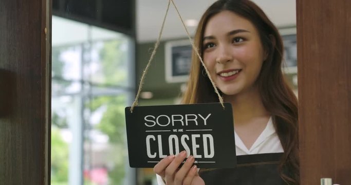 Close-up of a Attractive young asian woman turning over a "Open" sign in the morning at her cafe window and smiling looking outside waiting for clients.