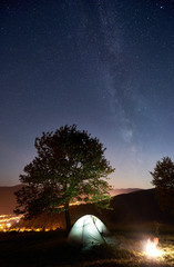 Tourist camping near big tree at summer night. Illuminated tent and campfire under amazing night sky full of stars and Milky way. On the background beautiful starry sky, mountains and luminous city