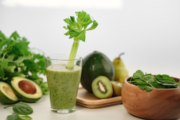 Green smoothie with a stalk of celery in a glass cup. On the table is an avocado.