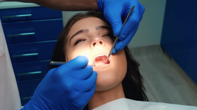young dentist wearing medical gloves, mask and glasses checks and cures patient woman's teeth using dental mirror and dam during appointment in dental clinic