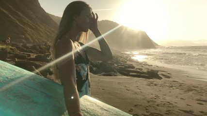 Young blonde surfer woman wearing colorful swimsuit combing her hair while is looking the sea with a blue surfboard on the other hand and sunset in background - Powered by Adobe