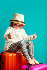 Joyful little girl is going to trip, sitting on suitcases, wearing a straw hat and sunglasses, shows thumbs up, portrait of cheerful child over isolated blue.