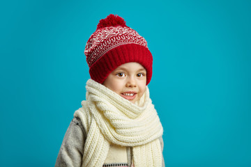 portrait of cute little girl in winter red hat and scarf on blue background, has a serious face, expresses surprise and shock