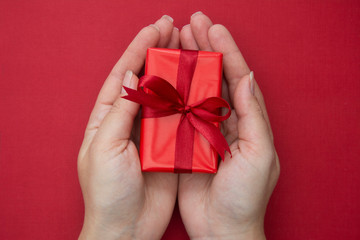 Female hands holding red Christmas gift box with red ribbon and bow, over red background. Flat lay.