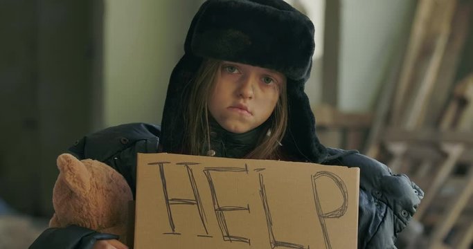 Portrait of sad homeless child holding the Help cardboard and looking at the camera. Little girl in hat with earflaps standing with a teddy bear and waiting for some help.