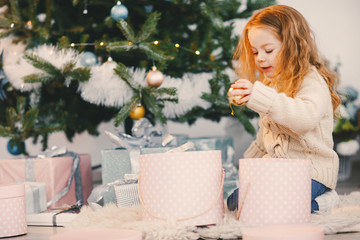 little blonde baby girl helping decorate the christmas tree by picking ornaments from a decor box and hanging them on the tree