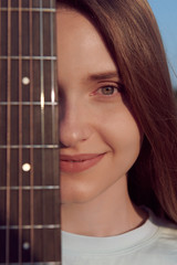 Closeup of a beautiful smiling girls face with guitar neck. Young musician with musical instrument. Caucasian girl with gorgeous eyes. Sunny day. Vertical photo.