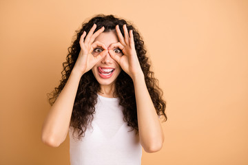 Fototapeta premium Closeup photo of funny lady playful mood holding round fingers in specs shape near eyes sticking tongue wear white casual outfit isolated beige pastel color background