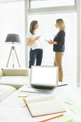 Fototapeta premium Blank screen of the laptop on the table at modern office. Female colleagues chatting on background while standing by the window at office