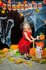 Fototapeta premium A child, a little girl in the shape of a witch on a broomstick, poses against the backdrop of scenery of cobwebs, pumpkins and autumn leaves on a Halloween holiday.