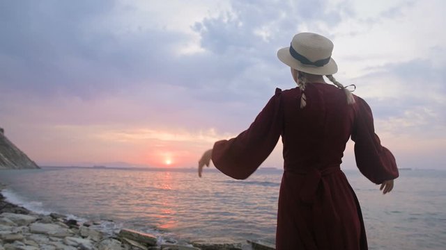 The view from the back. Slow motion Portrait of a young girl in a red dress and a straw hat on the seashore against a rocky shore and pink sunset.. Waiting for the return of the sailors