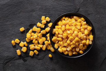 Canned sweet corn in a black ceramic bowl isolated on black slate next to spilled sweet corn. Top...